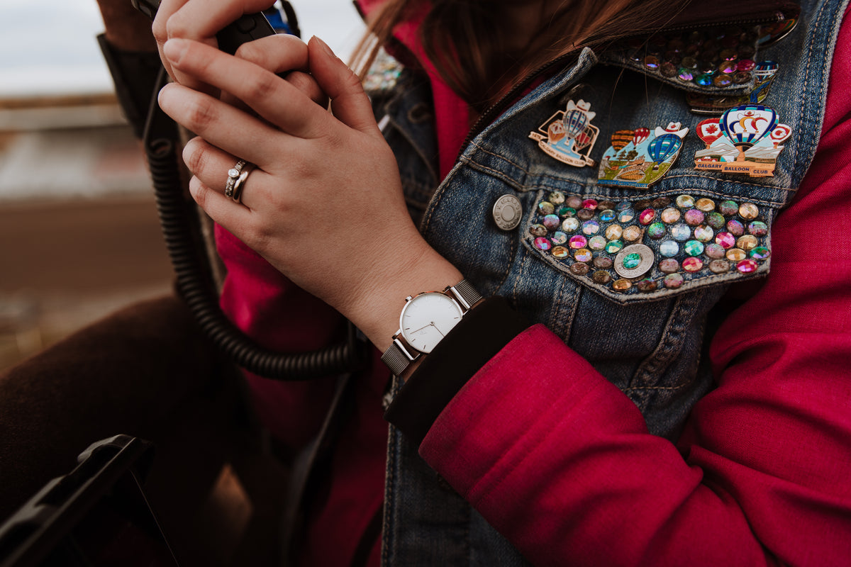 A MEDIUM watch with a white dial and silver mesh bracelet, worn with a red jacket and a denim vest covered in Calgary Balloon Club pins